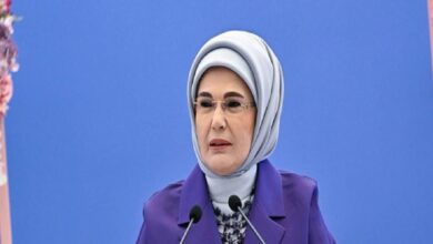 Middle-aged woman in a light gray hijab and purple blazer speaking at a podium with two microphones against a blue backdrop.