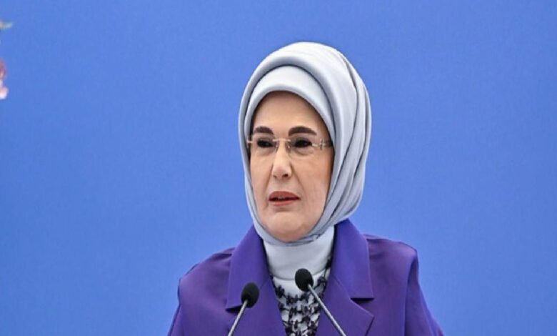 Middle-aged woman in a light gray hijab and purple blazer speaking at a podium with two microphones against a blue backdrop.