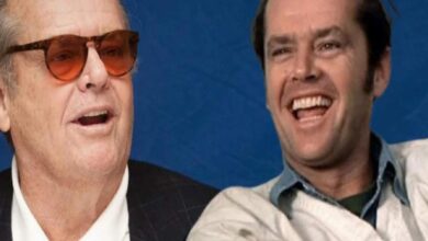 Two men laughing together against a blue backdrop; one wears sunglasses and a suit, the other in a light jacket.