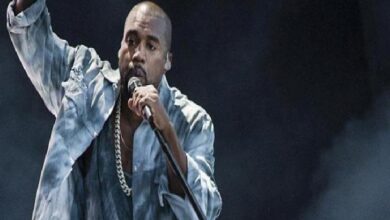 Male performer on stage, singing into a microphone with his arm raised, wearing a light denim jacket and gold chain against a dark backdrop.