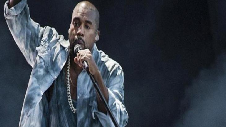 Male performer on stage, singing into a microphone with his arm raised, wearing a light denim jacket and gold chain against a dark backdrop.