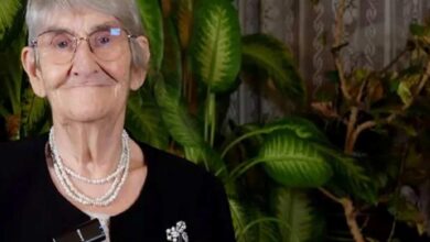 Elderly woman with short gray hair and glasses, wearing a layered pearl necklace and brooch, smiling with green houseplants in the background.