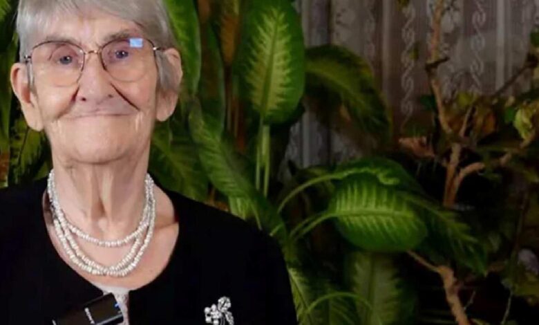 Elderly woman with short gray hair and glasses, wearing a layered pearl necklace and brooch, smiling with green houseplants in the background.