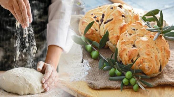 Hands kneading dough on a floured surface next to a rustic olive bread studded with olives and olive branches on a wooden board.