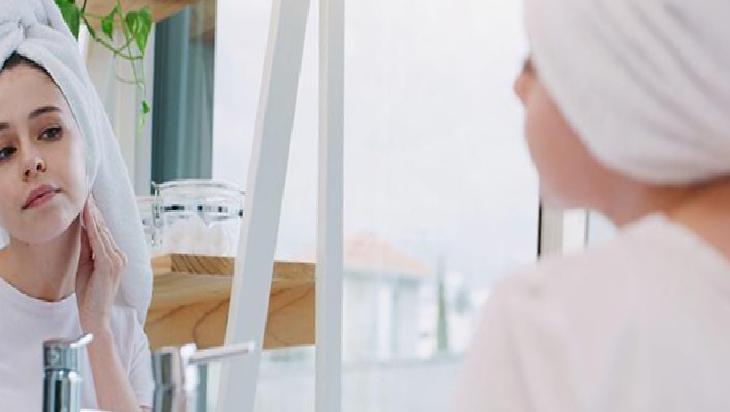Woman with towel on head looking at her reflection in a bathroom mirror, touching her jawline for skincare