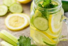 Glass mason jar of lemonade with cucumber, lemon slices and mint leaves, on a wooden table with celery in the foreground