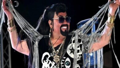 Stage performer wearing sunglasses and a black wig, adorned with multiple gold chains and silver garlands, smiling as they perform.