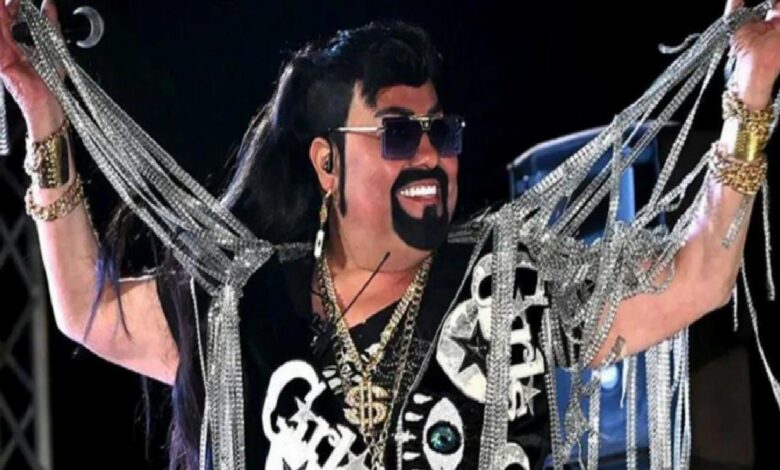 Stage performer wearing sunglasses and a black wig, adorned with multiple gold chains and silver garlands, smiling as they perform.