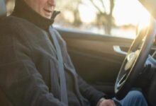 Person wearing a seatbelt and jacket, sitting in the driver's seat of a car with sunlight streaming through the window, ready to drive