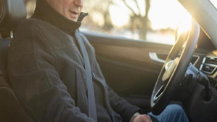 Person wearing a seatbelt and jacket, sitting in the driver's seat of a car with sunlight streaming through the window, ready to drive