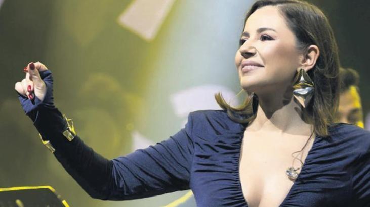 Female singer on stage with one arm raised, wearing a navy dress and large earrings, performing under bright stage lights.