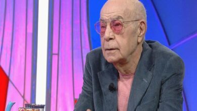 Older man with pink-tinted glasses and a pink shirt sits at a TV studio desk wearing a gray blazer and a lapel mic.