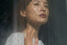 Woman gazing upward through a rain-soaked window, hand pressed against the glass.