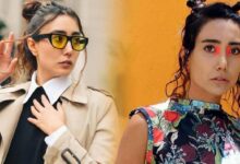 Two women posing outdoors: left in a beige trench coat with yellow sunglasses, right in a colorful floral top with red eye makeup.