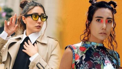 Two women posing outdoors: left in a beige trench coat with yellow sunglasses, right in a colorful floral top with red eye makeup.