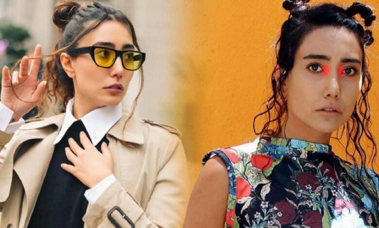 Two women posing outdoors: left in a beige trench coat with yellow sunglasses, right in a colorful floral top with red eye makeup.