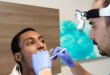 Dentist in a headlamp and blue gloves examines a patient's open mouth with dental tools during a checkup or procedure.