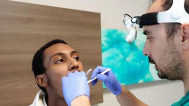 Dentist in a headlamp and blue gloves examines a patient's open mouth with dental tools during a checkup or procedure.