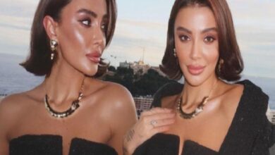 Two women in black off-shoulder outfits posing on a rooftop by the ocean, wearing statement necklaces and bold makeup as the sun sets behind them.