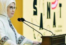 Woman in a white hijab and glasses speaks at a podium with two microphones at a formal event.