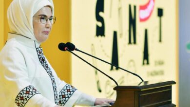 Woman in a white hijab and glasses speaks at a podium with two microphones at a formal event.
