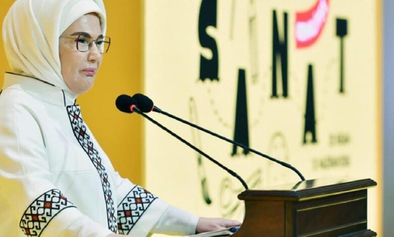 Woman in a white hijab and glasses speaks at a podium with two microphones at a formal event.