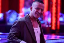 Smiling man in a brown blazer and white tank top leaning on a neon-lit bar in a nightclub setting.