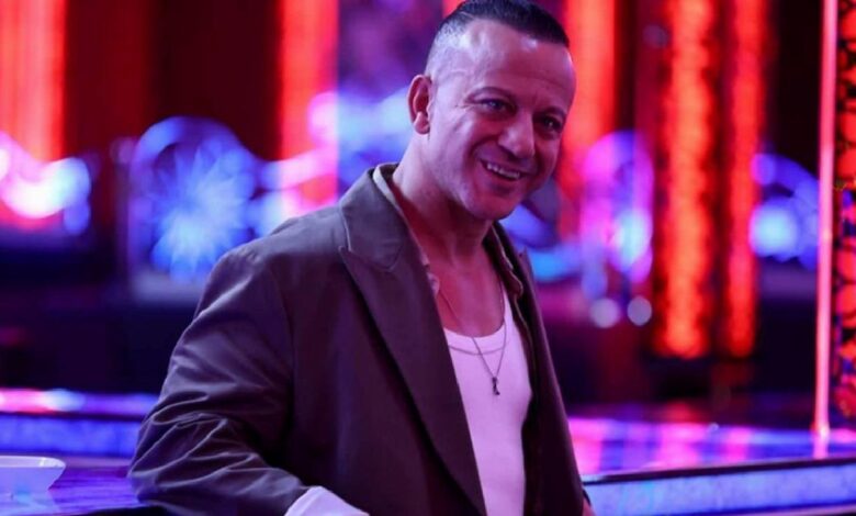 Smiling man in a brown blazer and white tank top leaning on a neon-lit bar in a nightclub setting.