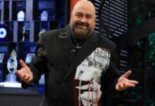 Smiling man in a black double-breasted blazer with a newspaper-print panel on the left, gesturing on a television studio set.