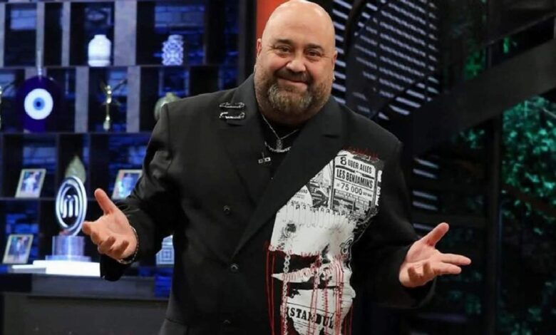 Smiling man in a black double-breasted blazer with a newspaper-print panel on the left, gesturing on a television studio set.
