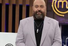 Bearded, bald man in a light gray blazer and black shirt on a TV studio set with a MasterChef logo in the background.
