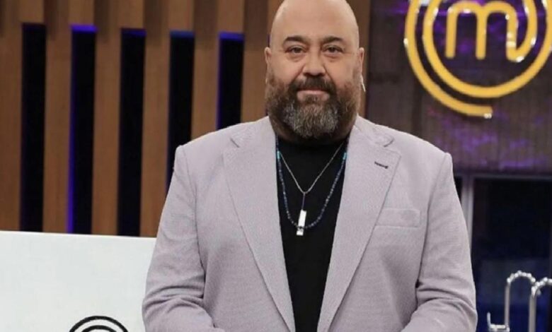 Bearded, bald man in a light gray blazer and black shirt on a TV studio set with a MasterChef logo in the background.