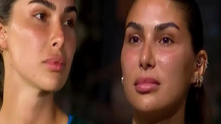 Two women with dark hair and glowing skin sit side by side, looking off to the left with serious expressions in a dim setting.