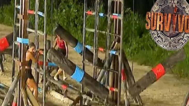 Survivor challenge on a sandy beach with tall wooden frames wrapped in blue and red tape and contestants in swimsuits, Survivor logo in the top right.