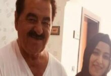 Two people pose indoors: a smiling man with a mustache in a white shirt and a woman in a dark hijab.