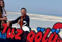 Two people stand beside a bright, cartoon-style sign on a salt flat beach, with a white flamingo illustration and red and blue lettering behind them.