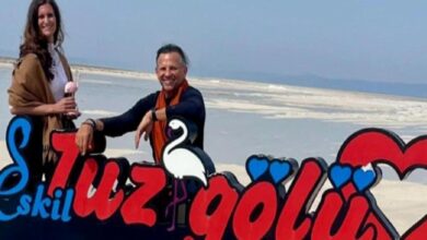 Two people stand beside a bright, cartoon-style sign on a salt flat beach, with a white flamingo illustration and red and blue lettering behind them.