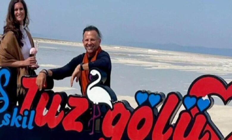 Two people stand beside a bright, cartoon-style sign on a salt flat beach, with a white flamingo illustration and red and blue lettering behind them.