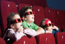 Three children wearing red 3D glasses sit in a cinema row, leaning on red seats and watching a screen.