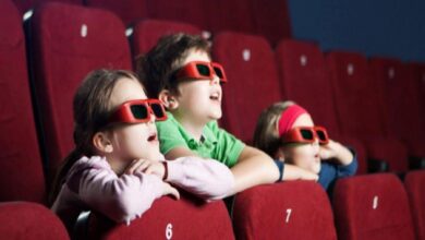 Three children wearing red 3D glasses sit in a cinema row, leaning on red seats and watching a screen.