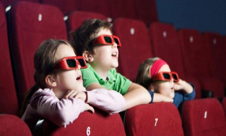 Three children wearing red 3D glasses sit in a cinema row, leaning on red seats and watching a screen.