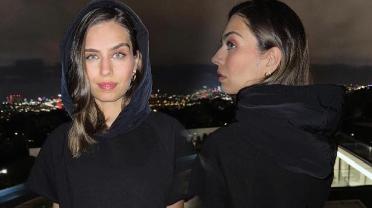 Two women in black hoodies on a rooftop balcony at night; one faces the camera while the other is in profile with city lights in the background.
