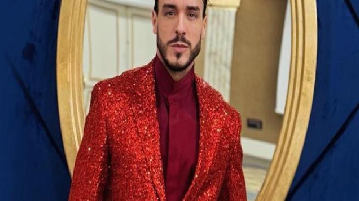 Man in a red glitter blazer and burgundy shirt posing indoors with a gold circular frame behind him.