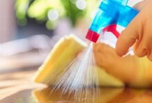 Hands hold a blue spray bottle and sweep a yellow sponge across a wooden surface for cleaning.