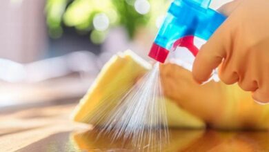 Hands hold a blue spray bottle and sweep a yellow sponge across a wooden surface for cleaning.