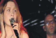 Female singer with red-tinted hair singing into a microphone on stage, a man in glasses blurred in the background.