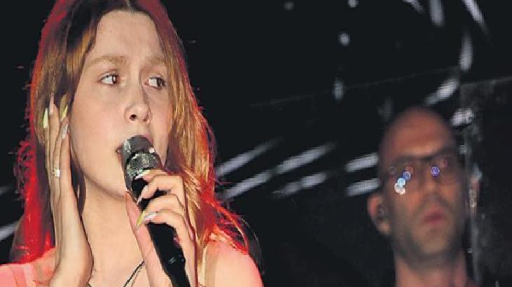 Female singer with red-tinted hair singing into a microphone on stage, a man in glasses blurred in the background.