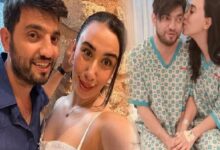 Selfie of a smiling couple in the foreground, casual attire, posing indoors beside a stone wall.