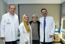 Four people in a modern exam room: two doctors in white coats with ID badges flank a smiling patient and a woman in a leopard-print top, all posing for a photo.