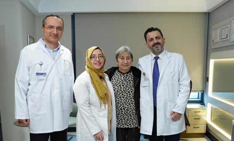 Four people in a modern exam room: two doctors in white coats with ID badges flank a smiling patient and a woman in a leopard-print top, all posing for a photo.